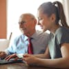 Young woman in casual clothes helping senior man in formal shirt with paying credit card in Internet using laptop while sitting at table